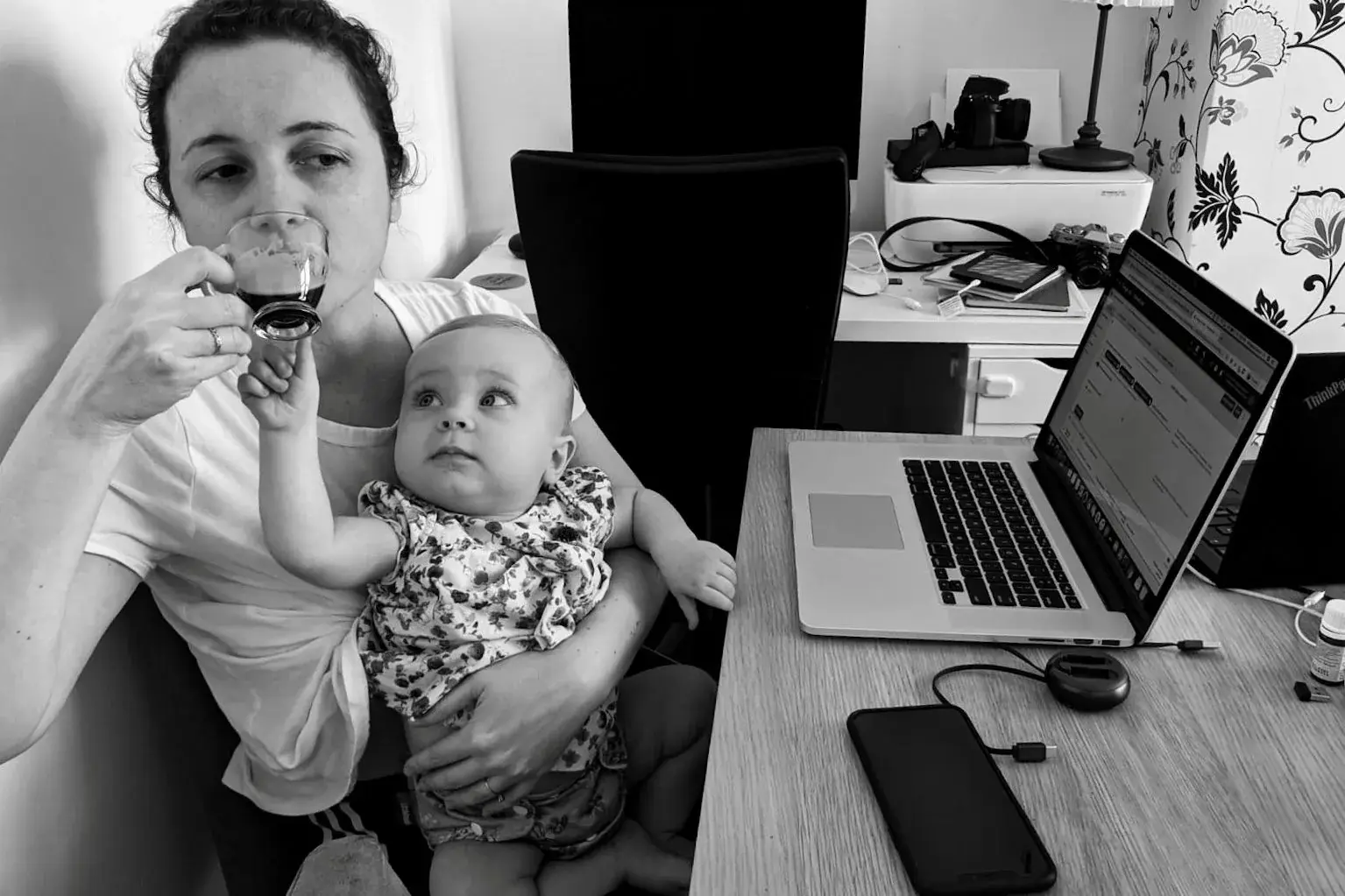 Mãe segurando um bebê no colo enquanto toma café ao lado de um notebook aberto em uma mesa de trabalho.